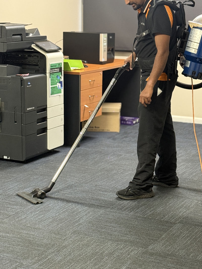 Cleaner vacuuming office carpet beside a photocopier with a backpack vacuum