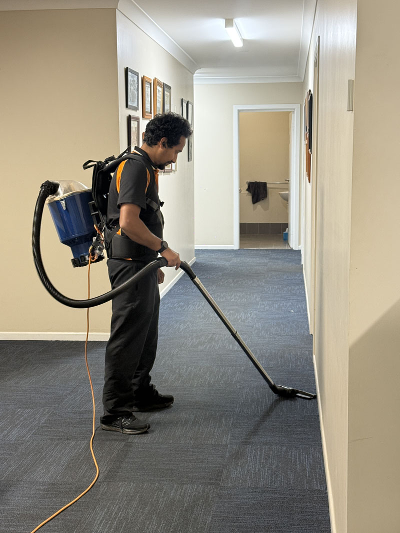 Cleaner with a backpack vacuum reaching further along a carpeted office hallway