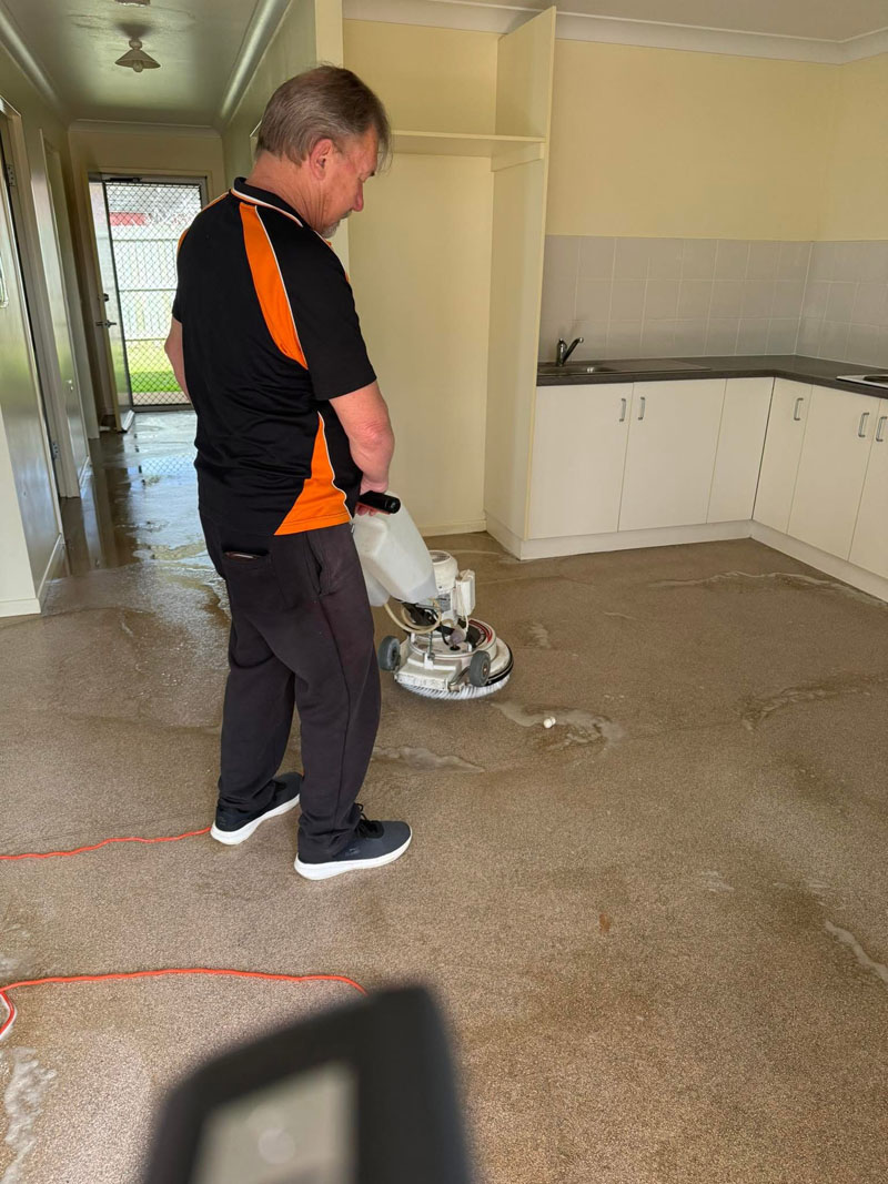 Cleaner scrubbing a wet vinyl floor with a floor machine near a kitchen