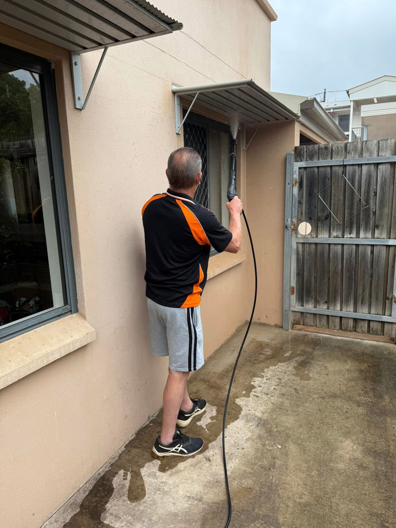 Cleaner hosing down an outside window and wall beside a wet concrete path