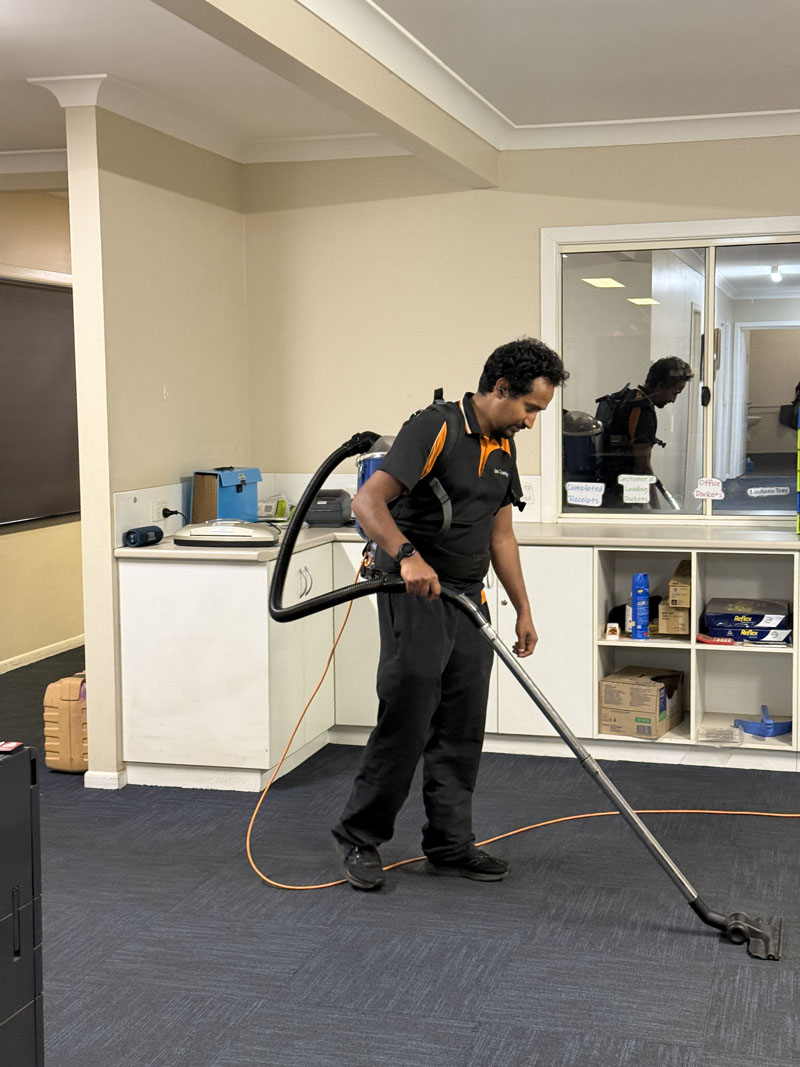 Cleaner vacuuming office carpet beside a kitchenette and shelving